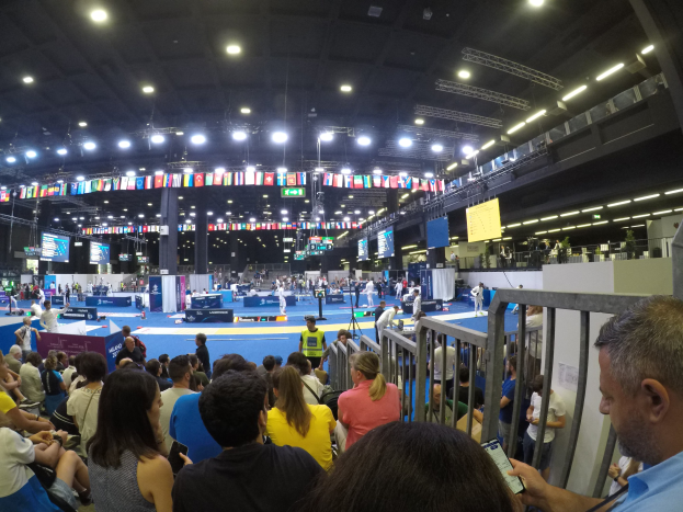 Eine große Menge von Menschen, die auf Sitzplätzen und Stehplätzen in einer Indoor-Sportarena sitzen, mit Bildschirmen, Flaggen und Deckenleuchten im Hintergrund.