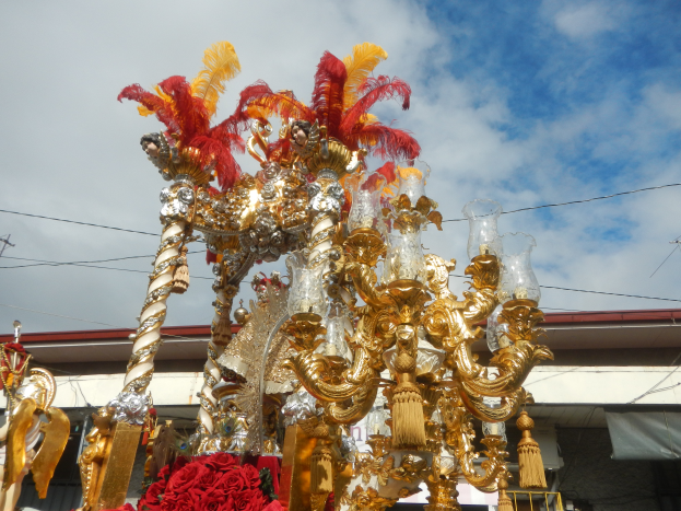 Ein prunkvoller gold- und roter Festwagen, geschmückt mit Blumen und anderen Dekorationen, in einem Karnevalsumzug, mit einem Gebäude, Strommasten mit Kabeln und einem bewölkten Himmel im Hintergrund.