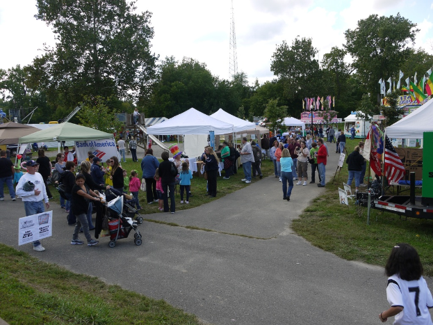 Eine Gruppe von Menschen, StÃ¼hle, Fahnen, BabystÃ¼hle, Zelte und eine Tafel sind auf einer grasigen FlÃ¤che mit BÃ¤umen im Hintergrund aufgebaut.