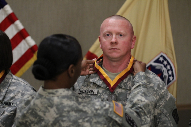 Eine Frau berührt die Medaille eines Mannes, der vor ihr steht, mit sichtbaren Fahnen im Hintergrund.