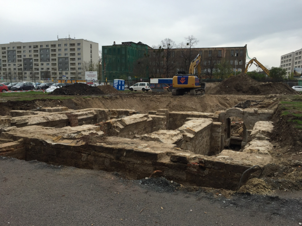 Eine Baustelle mit einem großen Haufen Erde, umgeben von Baggern, Fahrzeugen, Gras, Bäumen und Gebäuden im Hintergrund unter einem bewölkten Himmel, mit einer Straße unten.