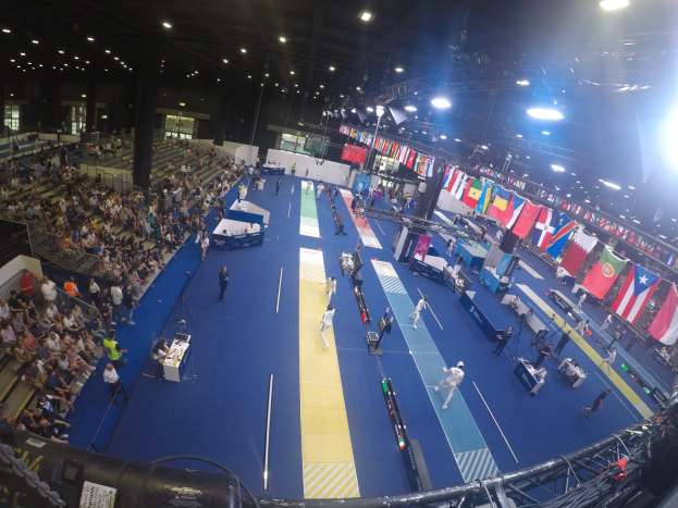 Großer Hallenstadium mit Zuschauern bei einer Gymnastikveranstaltung, auf beiden Seiten sitzend und einige stehend; Deckenleuchten und Flaggen im Hintergrund.