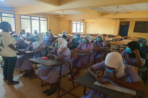 Ein Klassenraum in Nigeria mit Schülerinnen und Schülern, die Gesichtsmasken tragen und auf Bänken sitzen oder stehen, mit Büchern und Gegenständen auf den Tischen, Fenstern, einer Tür und Deckenleuchten und -ventilatoren im Hintergrund.