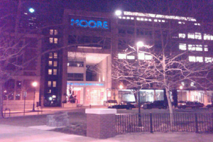 Ein Krankenhausgebäude mit mehreren Fahrzeugen, Bäumen und einem Zaun davor.