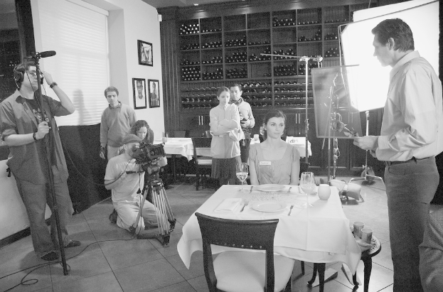 Ein Filmset mit einem Mann auf der rechten Seite, einer Frau, die an einem Tisch mit Tellern, Gabeln und Gläsern in der Mitte sitzt, und zwei Personen, die eine Kamera auf der linken Seite bedienen, mit Gestellen, Rahmen und einem Bildschirm im Hintergrund.