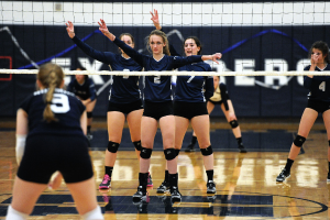 Drei Mädels auf einem Volleyballfeld mit erhobenen Händen, ein Netz davor und ein Zaun im Hintergrund.