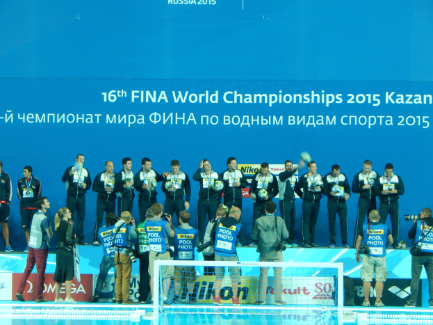 Eine Gruppe von Menschen steht oben auf einem Schwimmbad, einige halten Kameras, mit einem Netz im Vordergrund und einem Banner im Hintergrund, auf dem steht "FINA World Championships 2015 Kazan".