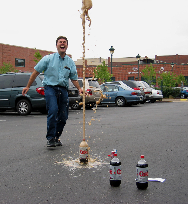 Ein Mann in einem blauen Hemd und Jeans steht lachend auf einer Straße vor der Stadt, mit drei leeren Flaschen in der Nähe, acht geparkten Autos, Bäumen, einer Straßenlaterne und einem roten Backsteingebäude im Hintergrund.
