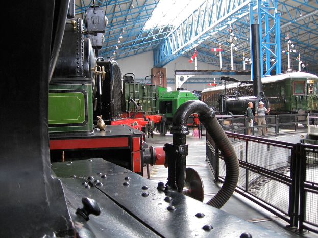 Mehrere Zugmaschinen stehen auf einem Abstellgleis mit zahlreichen Eisenstangen darüber. Rechts verläuft ein Zaun entlang einer Bahnstrecke mit einem Zug. Zwei Personen stehen in der Mitte der Szene in der Nähe des Zauns.