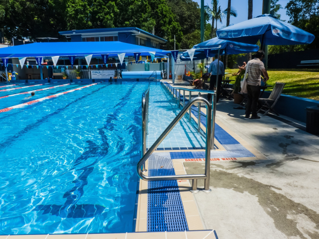 Großer Freibad mit schwimmenden Menschen, umgeben von Geländern, Sonnenschirmen, Stühlen, Tischen, Pflanzen, Gras, Bäumen und einem Gebäude unter einem klaren blauen Himmel.