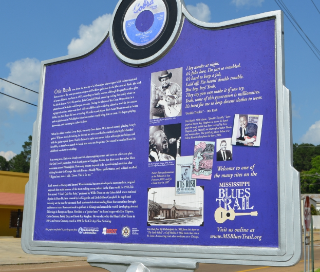 Ein Infotafel, Gebäude, ein Schuppen, Bäume, Stromkabel und ein bewölktes Himmel.