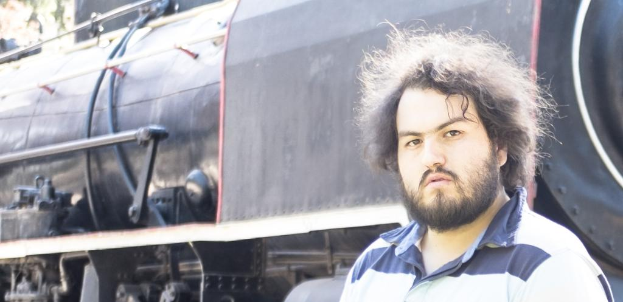 Ein Mann in einem T-Shirt steht neben einem Zug, wobei die Räder und andere Teile des Zugs unten und ein Baum oben links zu sehen sind.