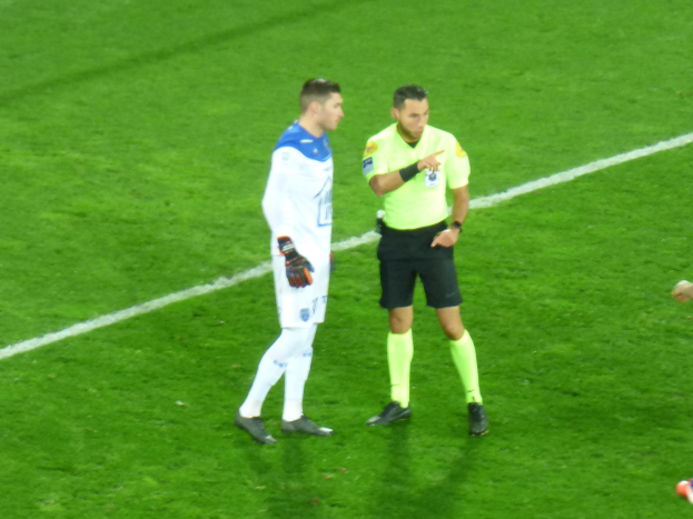 Ein Schiedsrichter in einem gelben Hemd und schwarzen Shorts spricht mit einem Fußballspieler in einem weißen und blauen Trikot auf dem Feld, beide tragen Turnschuhe.