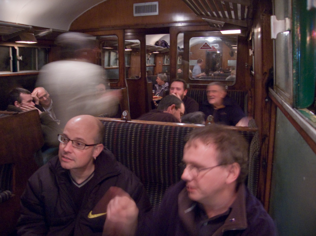 Zwei Männer sitzen auf einem Sitz in einem Abteil, mit einer Gruppe von Menschen, die dahinter sitzen, einer Tür zwischen den Abteilen oben und einem Fenster rechts.