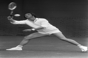 Ein Mann in einem wei├čem Tennisoutfit schwingt einen Schl├Ąger gegen einen Ball, mit entschlossener Miene und einer Wand im Hintergrund.