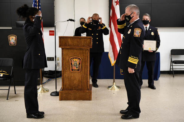 Eine Gruppe uniformierter Personen mit Masken bei einer Vereidigung eines neuen Feuerwehrchefs, mit einer Person, die ein Zertifikat hält, vor einem Podium mit Flaggen und sitzenden Zuschauern.