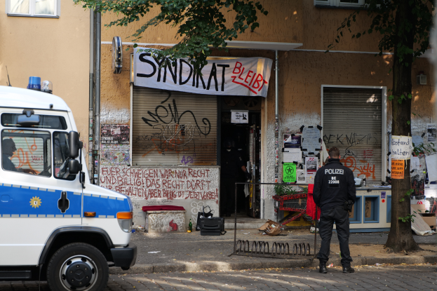 Ein Polizist steht vor einem Gebäude mit Graffiti, daneben ein Van, ein Baum und Gebäude mit Fenstern und einer Fahne im Hintergrund.