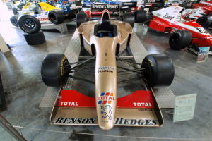 Rennwagen in einem Motorsportmuseum ausgestellt, getrennt von Besuchern durch einen Metallzaun, mit informativem Text im Hintergrund.