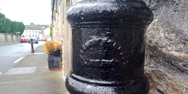 Ein schwarzer Bollard auf einer Straße neben einer Steinmauer, mit Fahrzeugen, Gebäuden, Bäumen und Polen im Hintergrund.
