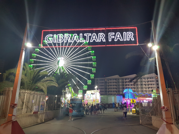 Der Eingang zur Gibraltar-Messe nachts, beleuchtet von einem großen Riesenrad und einer leuchtenden Schautafel, mit Menschenmassen, Bäumen und Gebäuden im Hintergrund unter einem dunklen Himmel.