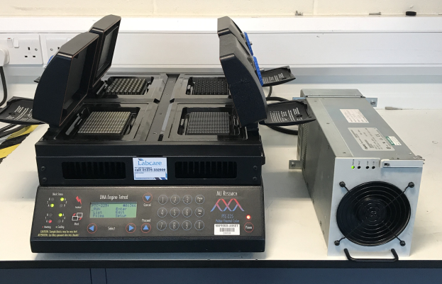 Zwei elektronische Geräte auf einem weißen Tisch in einem Laborumfeld, mit einer Schalttafel an der Wand im Hintergrund, wobei eines der Geräte ein DNA-Engine ist.