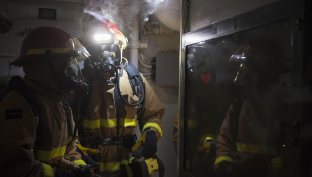 Feuerwehrleute in Gasmasken und Helmen stehen vor einer raucherfüllten Tür in einem beengten Raum mit Rohren im Hintergrund.