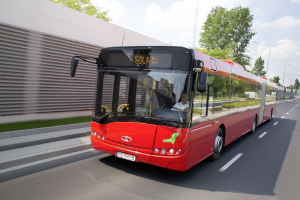 Ein roter Bus fährt auf einer Straße neben einem Gebäude mit einer Person im Inneren, neben einer Wand, Bäumen, Laternen und einem bewölkten Himmel im Hintergrund.
