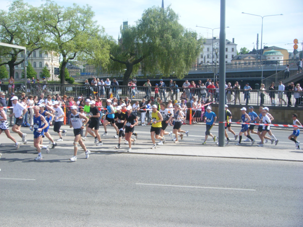 Eine Gruppe von Menschen, die bei einem Marathon auf einer Straße laufen, umgeben von einem Metallzaun, einem Tor, einem Band an einem Pfahl, Zuschauern auf dem Gehweg, einer Metallabsperrung, Pfählen, Straßenlaternen, Schildern, einer Brücke, Gebäuden mit Fenstern, Bäumen und einem bewölkten Himmel.
