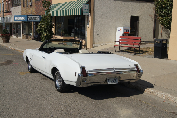 Ein weißes Auto mit der Nummer 771-299 steht auf der Straße, rechts daneben eine Bank, im Hintergrund ein Gebäude und ein Geschäft namens "Immobilienversicherung".