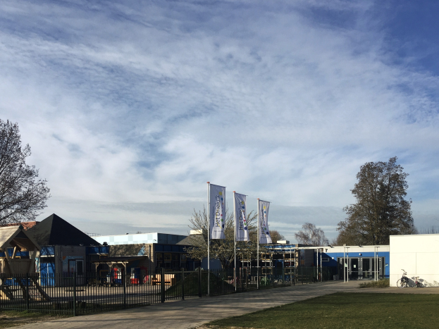 Großes modernes Schulgebäude mit einem grasigen Hof, der von einem Metallzaun umgeben ist, mit einem gepflasterten Weg, Bäumen, Bannern, einem geparkten Fahrrad, nahegelegenen Häusern und einem bewölkten Himmel.