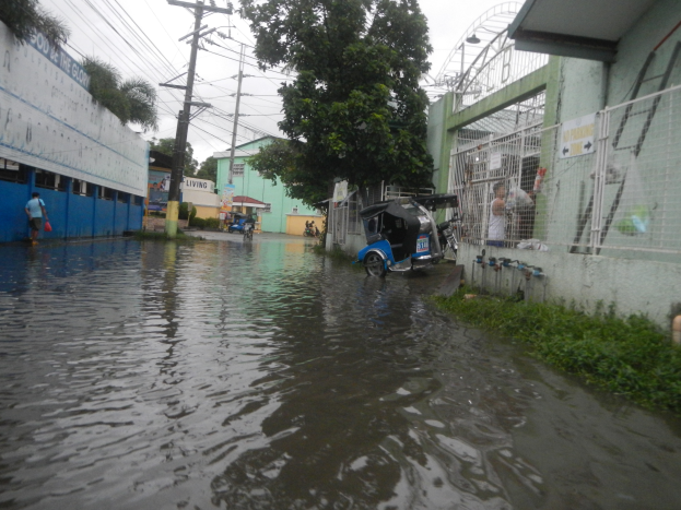 Eine �berflutete Stra�e mit einem geparkten Motorrad, Geb�uden, B�umen und Strommasten auf beiden Seiten sowie ein paar Menschen und Fahrzeugen im Hintergrund.