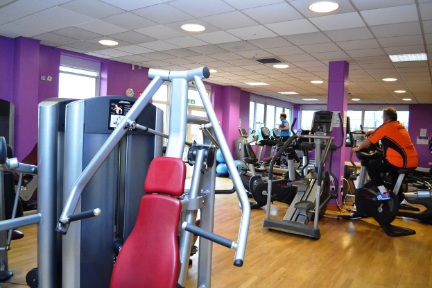 Ein Fitnessstudio mit mehreren Ger√§ten und ein paar Menschen beim Training, mit einer Decke und Lampen oben im Bild.