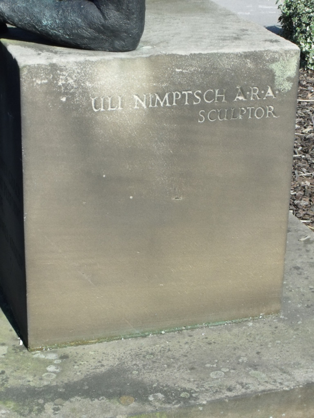 Ein Stein mit Text in Vordergrund, möglicherweise eine Statue oben, trockene Zweige, Erde und Pflanzen rechts, mit einer Straße im Hintergrund.
