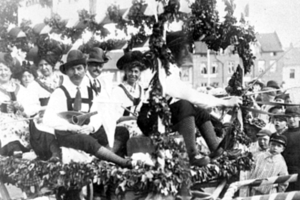 Eine Gruppe von Menschen in traditioneller bayrischer Tracht, einige mit Musikinstrumenten, fährt auf einem blattgeschmückten Festwagen in einem Umzug, mit Gebäuden und einem klaren Himmel im Hintergrund, dargestellt in Schwarz-Weiß.