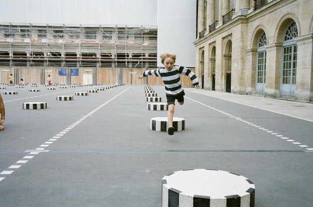 Ein Spielplatz mit vier Kindern, eines läuft auf dem Boden mit schwarzen und weißen Betonstufen, umgeben von einem Schulgebäude.