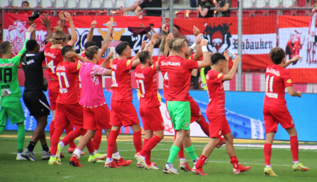 Gruppe von Fußballspielern, die ein Tor feiern, auf einem Feld mit Flaschen haltenden oder klatschenden Spielern, mit Bannern, einem Zaun, Metallstangen, Stühlen und einem Dach mit Lampen im Hintergrund.