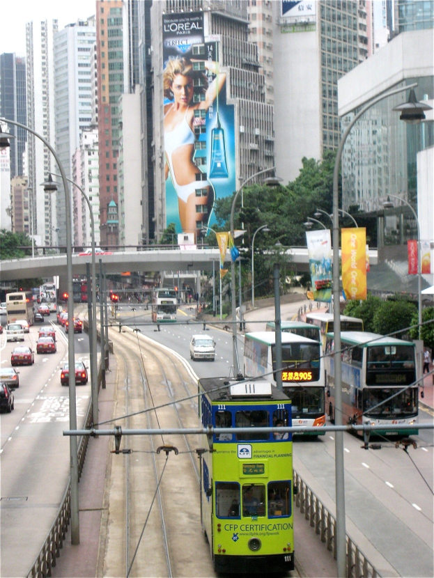 Eine belebte Stadtstra├če mit Doppeldeckerbussen, Autos und anderen Fahrzeugen unter einem klaren blauen Himmel, mit hohen Geb├Ąuden, Laternen, Schildern, B├Ąumen und einer ├ťberquerung im Hintergrund.