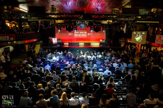 Eine große Menschenmenge sitzt auf Stühlen und schaut auf eine Bühne mit Bildschirmen und Plakaten, die von Scheinwerfern beleuchtet und von Geländern umgeben sind, mit einem Wasserzeichen in der linken unteren Ecke.
