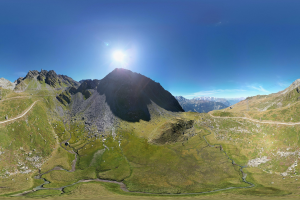 Panoramischer Blick auf ein Gebirgsmassiv mit grüner Wiese, einer gewundenen Straße und einem hellblauen Himmel mit Sonnenlicht.