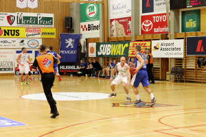Ein Basketballfeld mit Spielern im Vordergrund und einer hölzernen Wand mit Postern und Bannern im Hintergrund.
