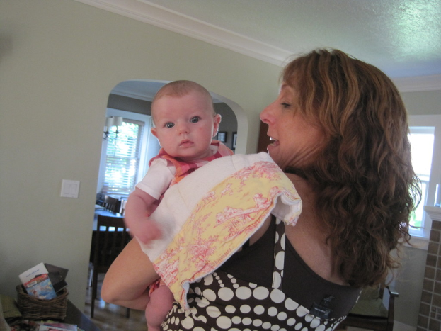 Eine Frau mit einem Baby auf dem Arm, das von einem Tuch auf ihren Schultern bedeckt ist, die in einem Raum mit einem Korb voller Bücher, einem Esstisch, Lampen, Fotorahmen und Fenstern im Hintergrund steht.