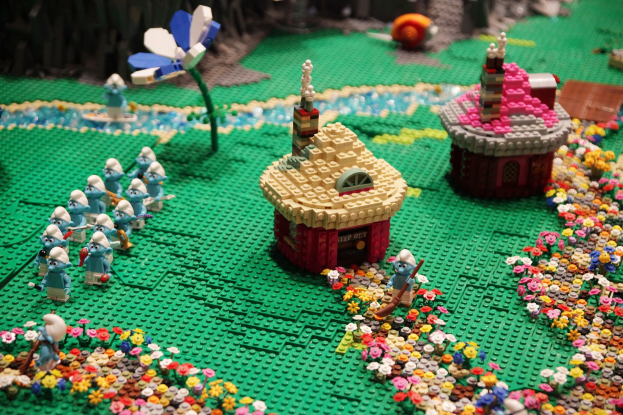 Ein großer Raum mit bunten LEGO-Bausteinen, die auf dem Boden verstreut sind, umgeben von Tischen und Säulen, mit Glasfenstern, die eine lebhafte Museumszenen draußen zeigen.