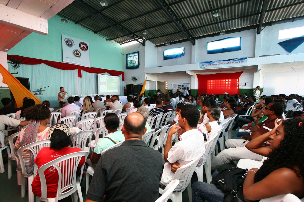 Eine Gruppe von Menschen sitzt auf Stühlen und schaut auf eine Bühne, auf der ein Polizeibeamter in ein Mikrofon spricht, wobei auf der rechten Seite des Bildes eine Tür zu sehen ist.