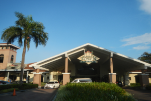 Der Eingang eines Resorts mit Palmen vorne, umgeben von Gebäuden, Fahrzeugen auf der Straße, Verkehrshütchen, Büschen, Bäumen und einem bewölkten Himmel im Hintergrund.