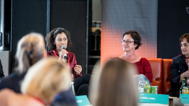 Eine Gruppe von Frauen sitzt in der ersten Reihe, wobei eine Frau links eine rote Kleidung trägt und ein Mikrofon hält, und im Hintergrund eine Rolltafel und eine schwarze Plane zu sehen sind.