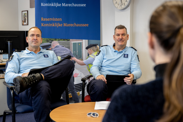 Zwei Polizeibeamte sitzen an einem Tisch mit einer Frau, umgeben von Dokumenten und Gegenständen, mit einem Banner, einer Uhr und Fotorahmen an der Wand und einem weiteren Tisch mit Computerausrüstung links daneben.