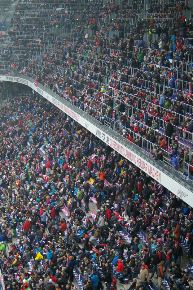 Eine große Menschenmenge sitzt und steht in einem Stadion mit Geländern und Texttafeln im Hintergrund.