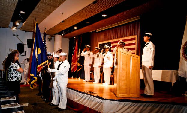 Eine formelle Veranstaltung mit Menschen, die stehen und salutieren auf einer Bühne, Flaggen in rot, blau und weiß, Stühle, ein Podium und ein Tisch mit einer weißen Decke, mit einem Fernseher und einem Lautsprecherbox im Hintergrund.
