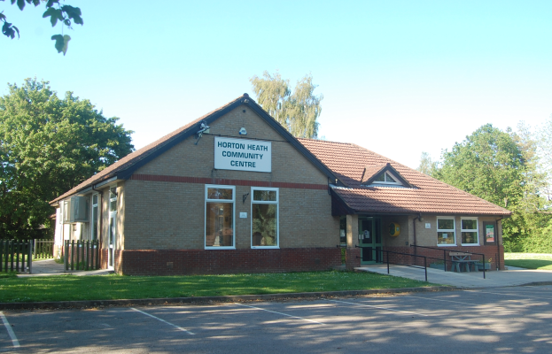 Ein Haus mit Dach, Fenstern und einem Schild mit der Aufschrift "Horton Heath Community Centre", umgeben von einem Metallzaun, einer Bank, Pflanzen, Gras, einem Weg, einer Baumgruppe und einem bewölkten Himmel.