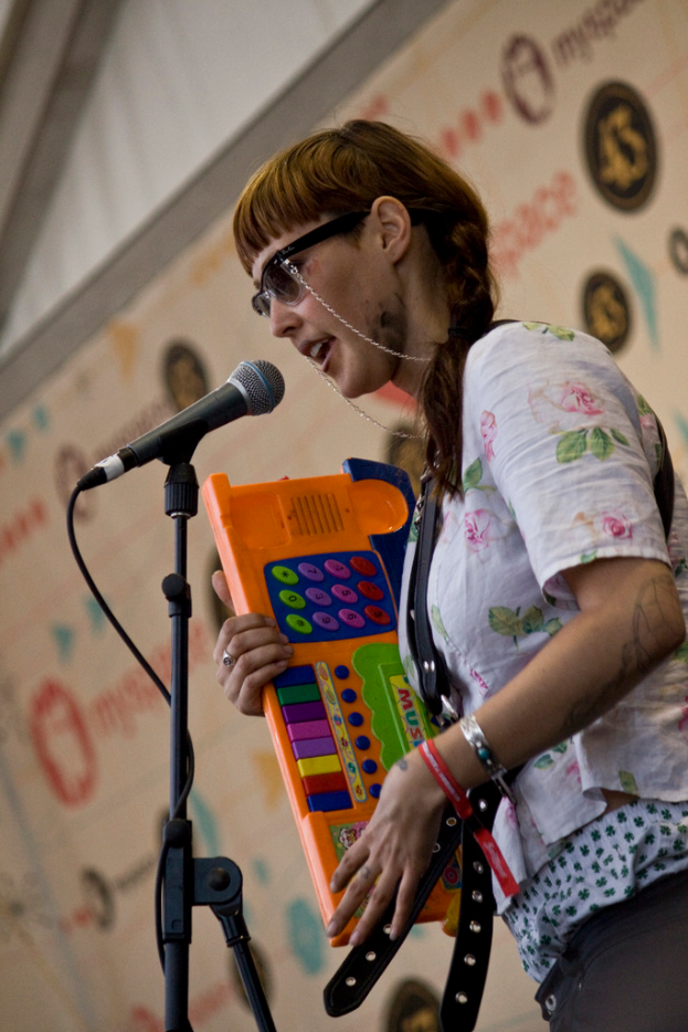 Eine Frau in einem weißen Hemd und Sonnenbrille singt in ein Mikrofon auf einem Ständer und hält dabei ein Piano, während ein Plakat an der Wand hinter ihr hängt.
